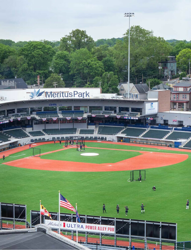 A view of an empty baseball stadium, Meritus Park, with a few people on the field, green grass, empty stands, and houses and trees in the background. American and Maryland flags are visible in the foreground.