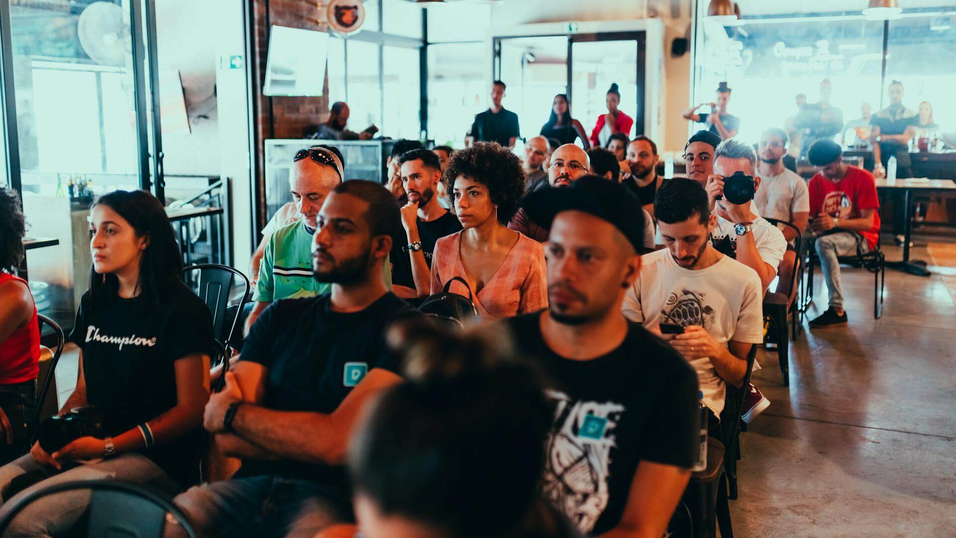 A diverse group of people sits attentively in rows of chairs at an indoor event or seminar, with some individuals focused forward and others looking at their phones. The setting is casual and well-lit.