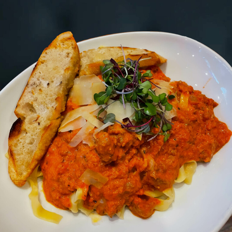 A white bowl of pasta topped with creamy tomato meat sauce, shaved parmesan, and microgreens, served with two slices of toasted bread.