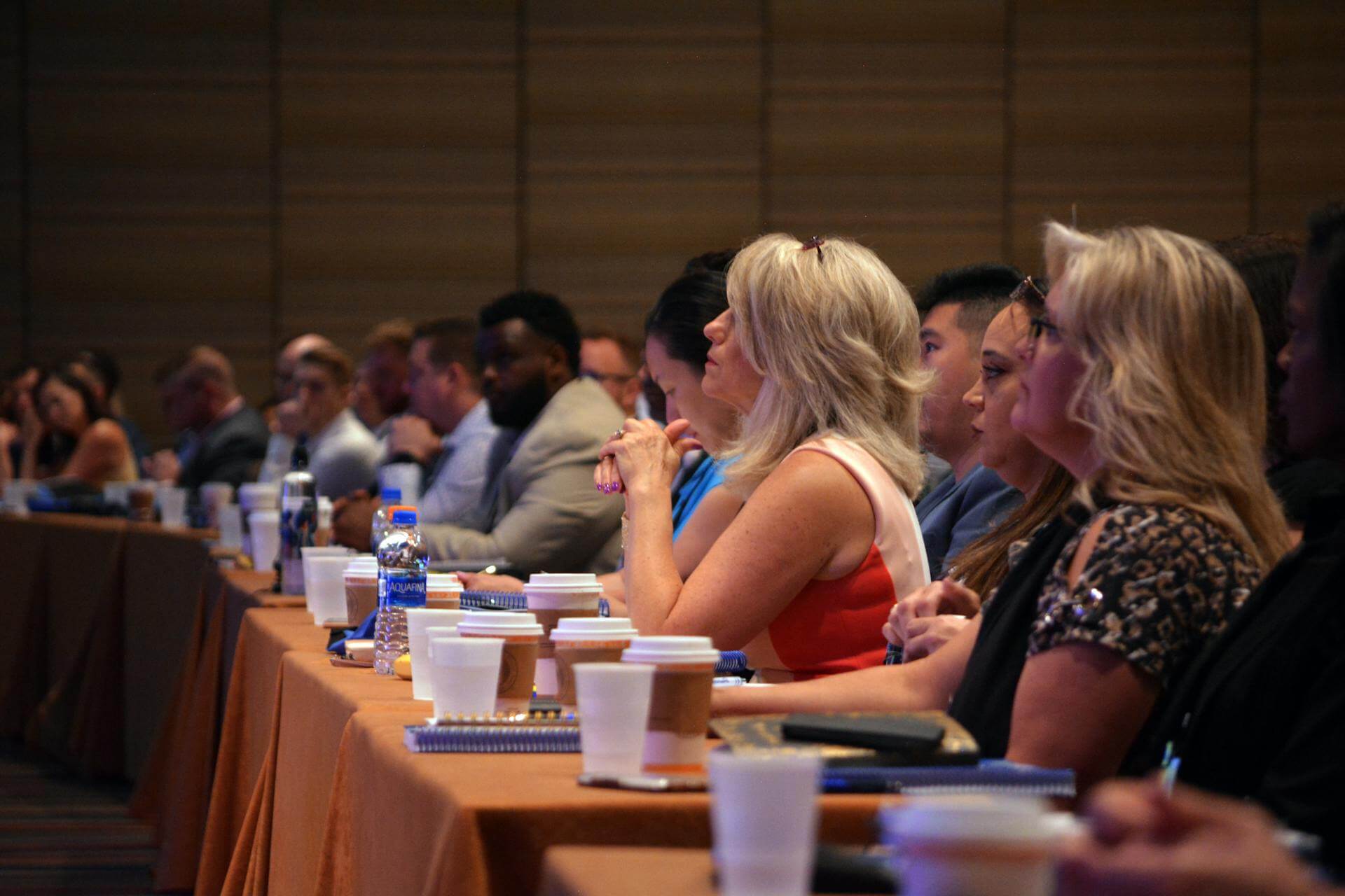 A group of people sit in rows at a conference or seminar, listening attentively. Notebooks, coffee cups, and water bottles are placed on the tables in front of them.