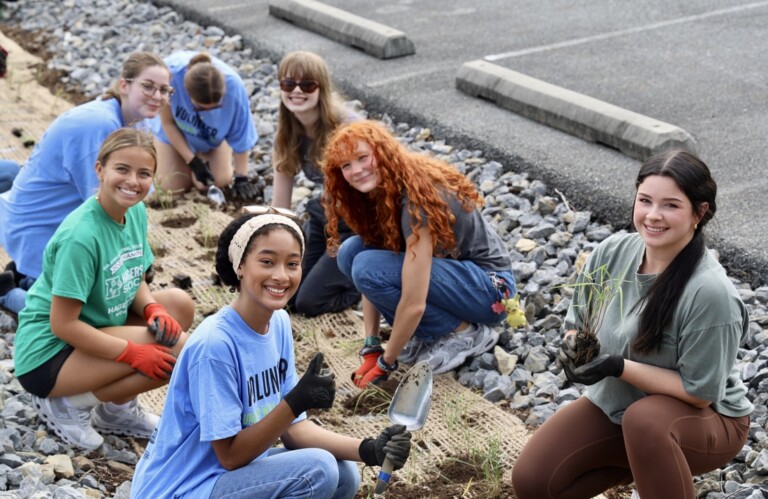 A group of young people kneel on rocky ground, planting small plants near a parking lot; they are smiling and wearing gloves and casual clothing.