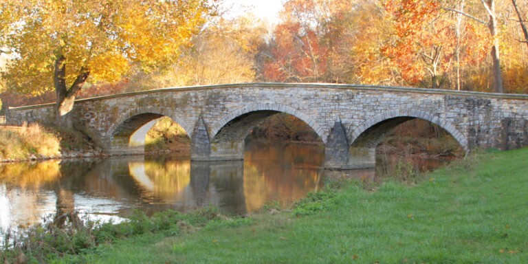 A stone arch bridge crosses a calm river, surrounded by autumn trees with orange and yellow leaves. The bridge and foliage are reflected in the water, creating a peaceful, scenic landscape.
