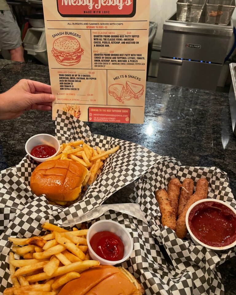 A hand holds a Messy Jessy’s menu above a counter with two trays of food, each featuring a burger, fries, and ketchup—plus mozzarella sticks with marinara sauce—at the cozy dog house–themed eatery.
