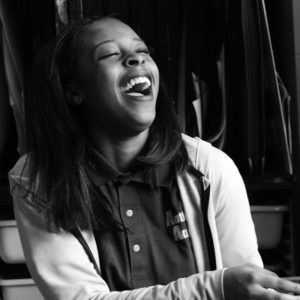A young woman wearing a collared shirt and jacket laughs joyfully, her head tilted back. The black and white photo captures her indoors, with shelves and hanging clothes behind her—perhaps pausing during an adventure on a cultural-trail.