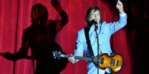 A musician in a light blue suit sings and plays guitar on stage in front of a red curtain, with a large shadow cast behind him.