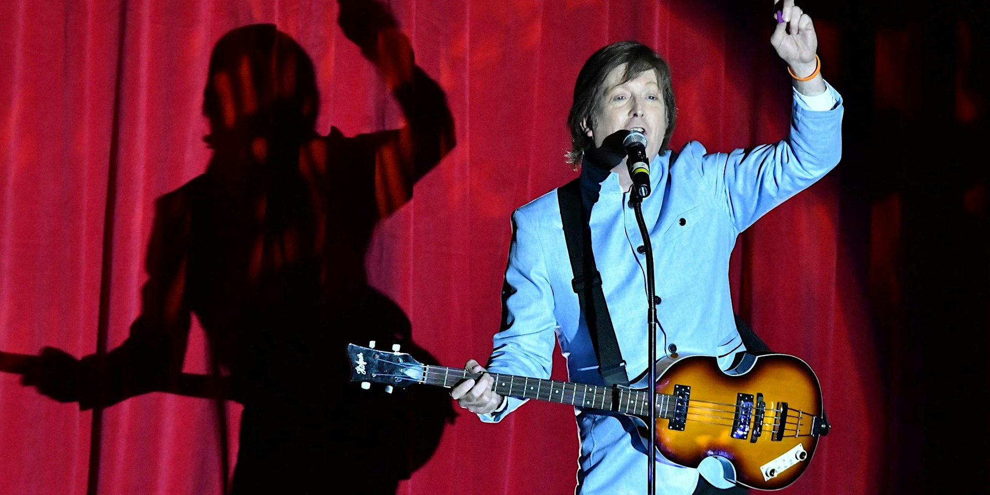 A musician in a light blue suit sings and plays guitar on stage in front of a red curtain, with a large shadow cast behind him.