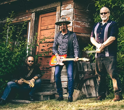 Three men with musical instruments pose outside an old wooden building. One sits holding a small box instrument, while the other two stand, one with an electric guitar. All wear sunglasses and casual clothes.