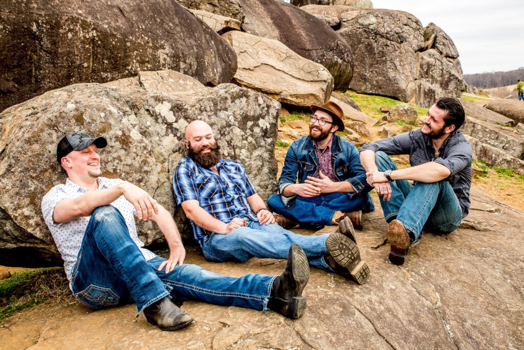 Four men sit on large rocks outdoors, laughing and talking together. They are casually dressed in jeans, shirts, and hats, with a natural rocky landscape in the background.