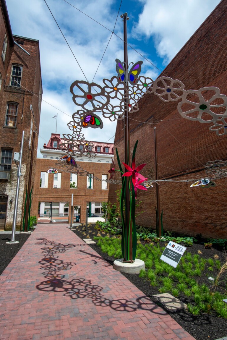 A brick walkway between tall brick buildings along the cultural-trail features a colorful outdoor art installation with large metal flowers and butterflies, casting intricate shadows on the path. There is a garden and a white information sign nearby.
