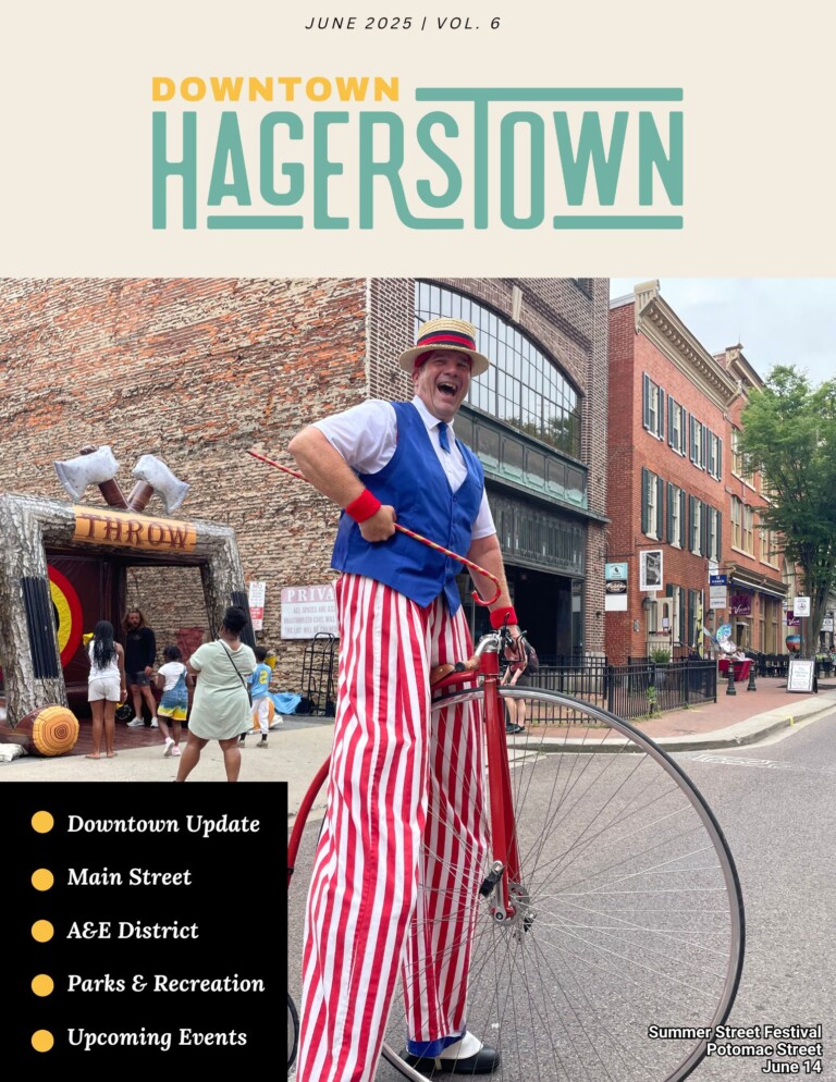 A man in a red bowler hat, blue vest, and red-and-white striped pants rides a vintage penny-farthing bicycle at a lively downtown street festival in Hagerstown, with people and historic buildings in the background.