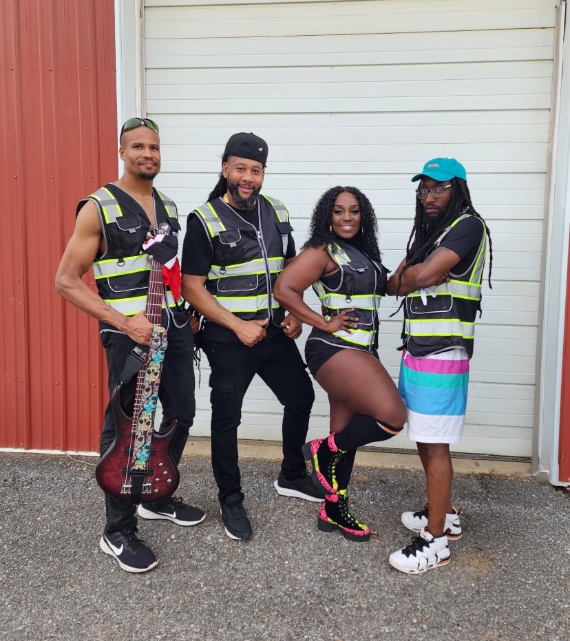 Four people pose confidently in front of a garage door, all wearing black outfits with reflective safety vests. One person holds a guitar, and another stands with one leg lifted, showing off colorful knee-high socks.