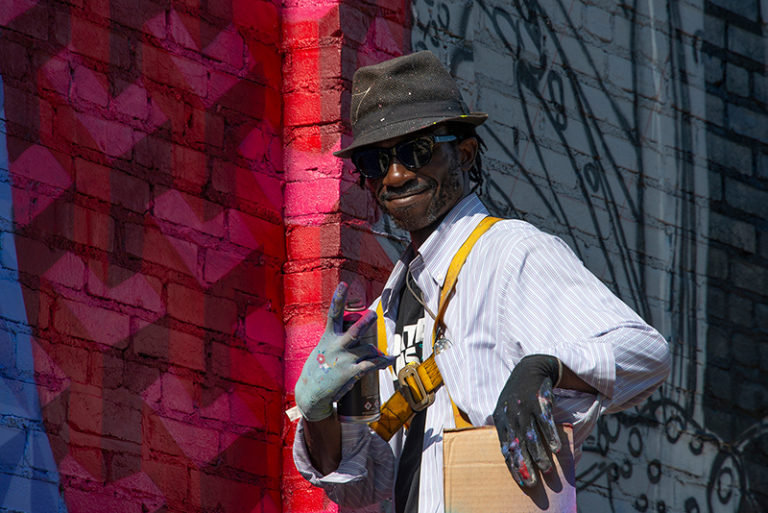 A smiling man wearing a hat, sunglasses, and gloves, with paint on his hands, stands in front of a colorful mural on a brick wall, making a hand gesture and holding a cardboard box.