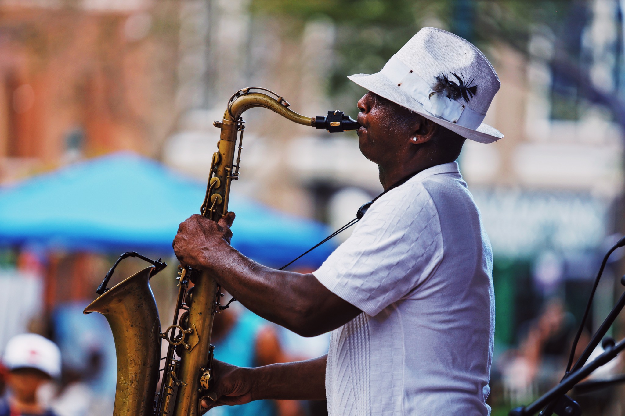 A man in a white hat with a feather and a white shirt plays a saxophone outdoors, with a blurred background of people and tents.