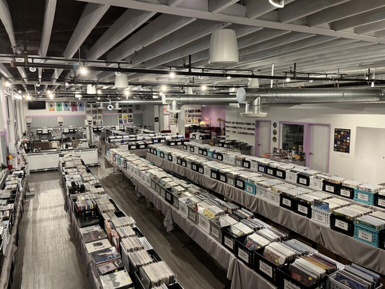 A spacious, well-lit record store interior with rows of bins filled with vinyl records. The room has exposed ceilings, light gray floors, and walls decorated with album covers and posters.