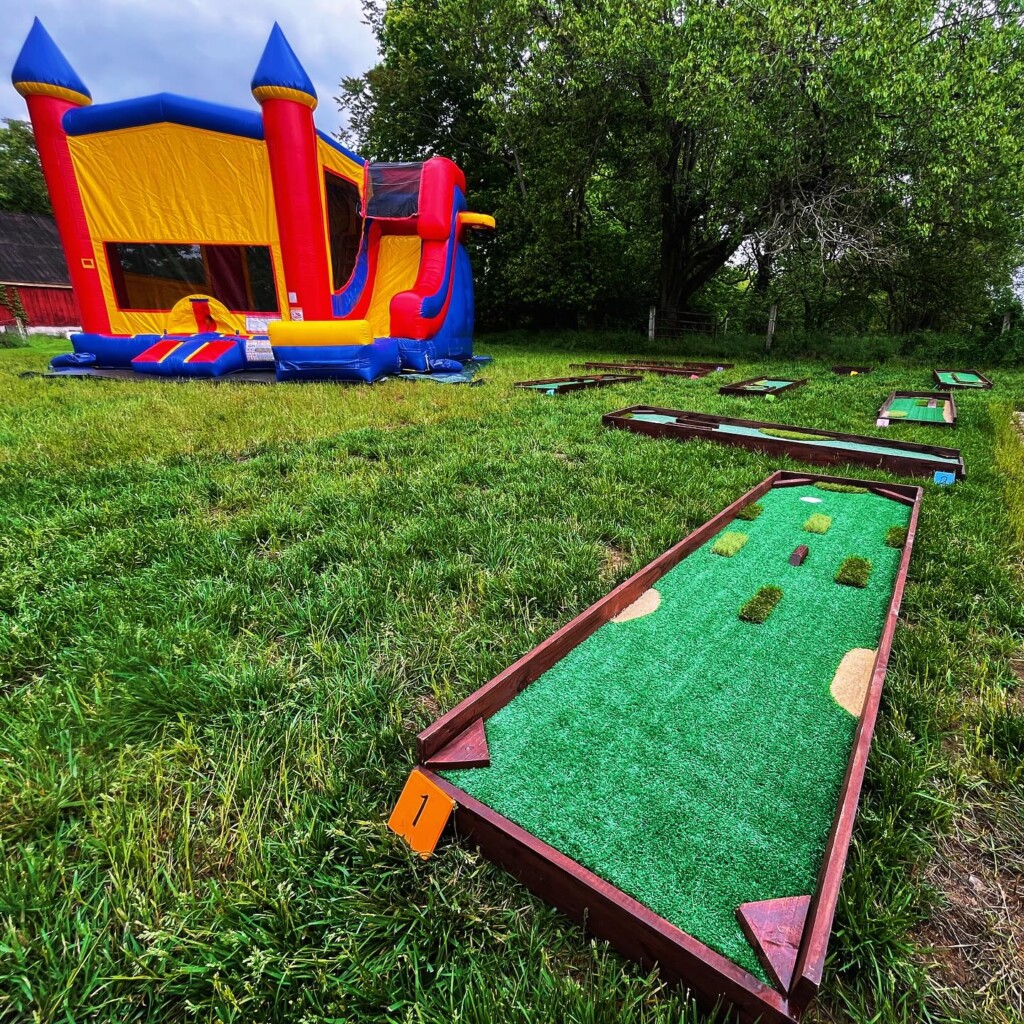 A colorful inflatable bounce house sits on grass beside several mini-golf holes with wooden borders and artificial turf, set up outdoors with trees in the background.