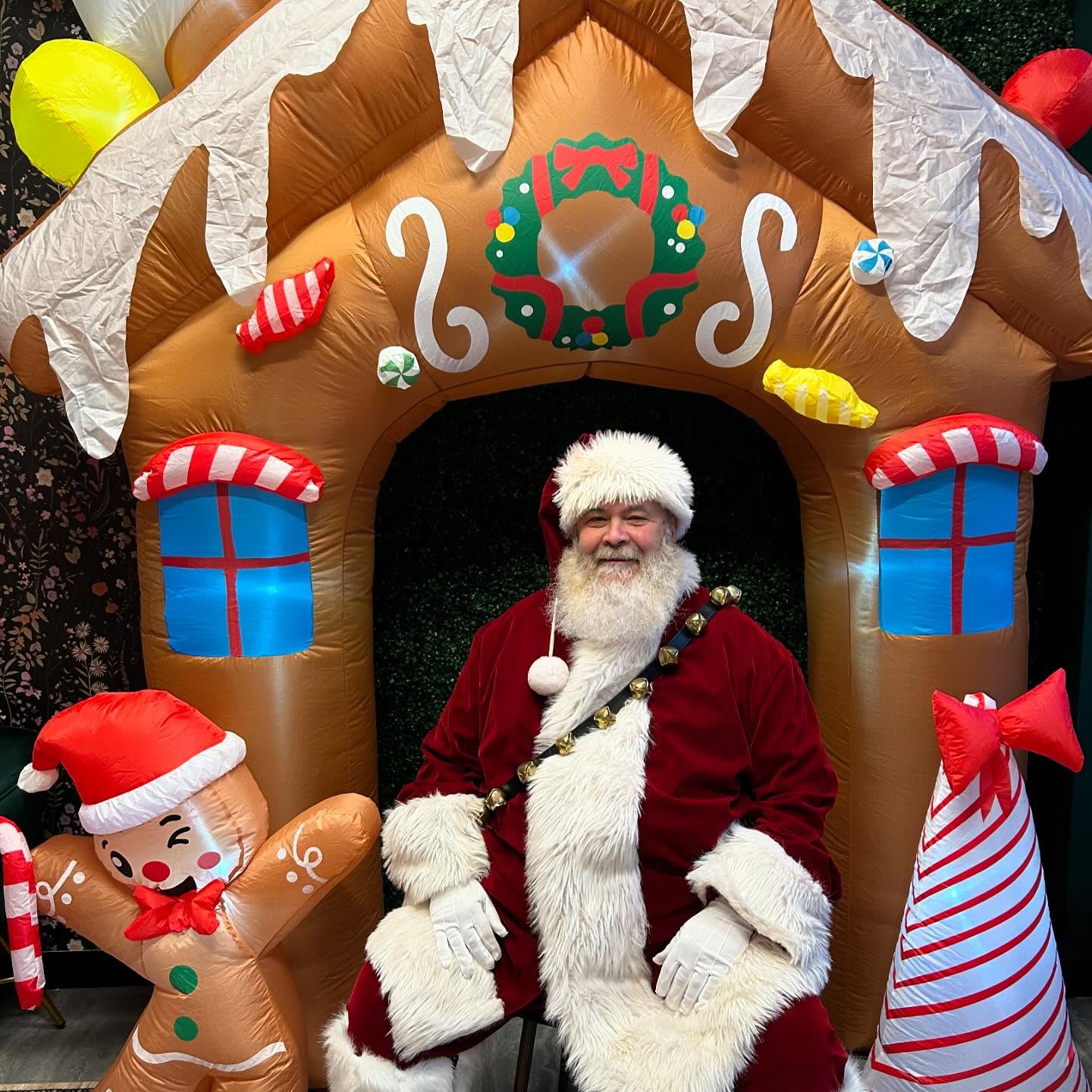 A man dressed as Santa Claus sits smiling in front of an inflatable gingerbread house, surrounded by festive decorations including a gingerbread figure and a red-and-white striped tree.