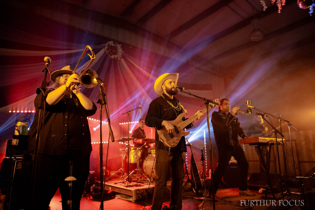 A live band performs on stage under warm, colorful lights. A trombone player, bassist in a cowboy hat, keyboardist, and drummer are visible. The venue has draped fabric and string lights. The atmosphere is lively and energetic.