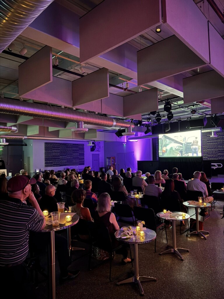 A group of people sit at small round tables with drinks, watching a large screen in a dimly lit, modern event space with purple lighting and industrial decor.