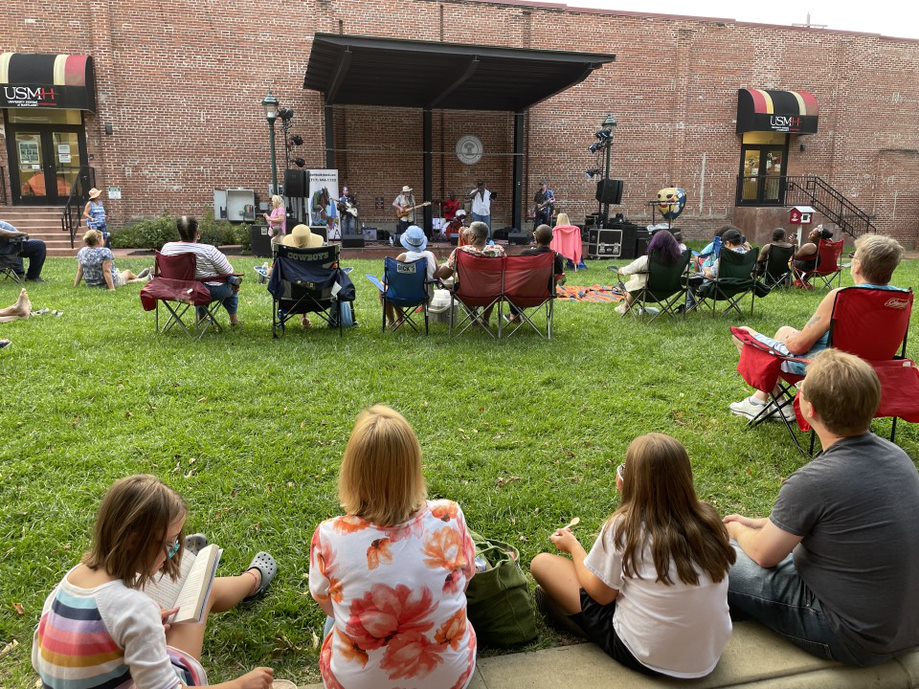 People sit on lawn chairs and blankets, enjoying a relaxed mdwk evening as a live band performs on a small outdoor stage in front of a brick building. Some audience members are reading or simply unwinding on the grass.