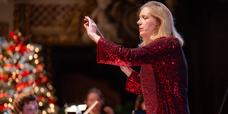 A woman in a sparkly red top conducts an orchestra, with a decorated Christmas tree blurred in the background. Musicians can be seen playing their instruments behind her.