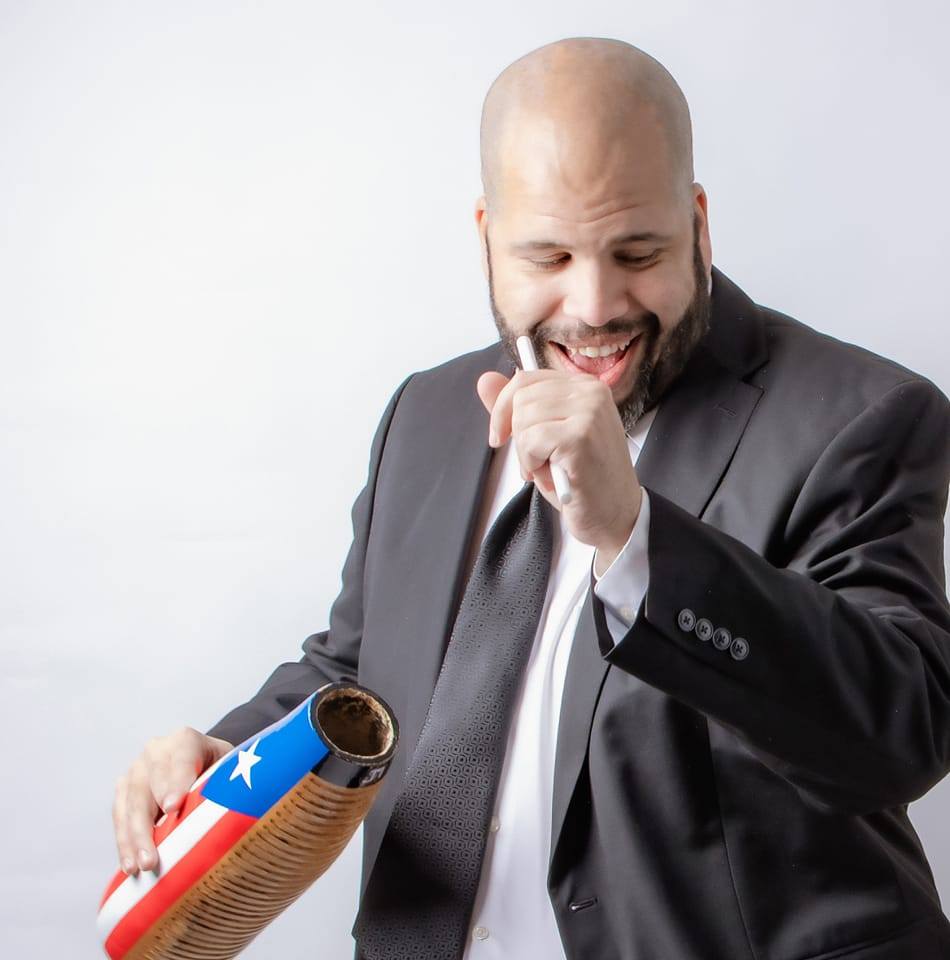 A man in a black suit and white shirt smiles while playing a güiro decorated with the Puerto Rican flag. He holds a scraper in one hand and looks down, appearing joyful against a plain white background.