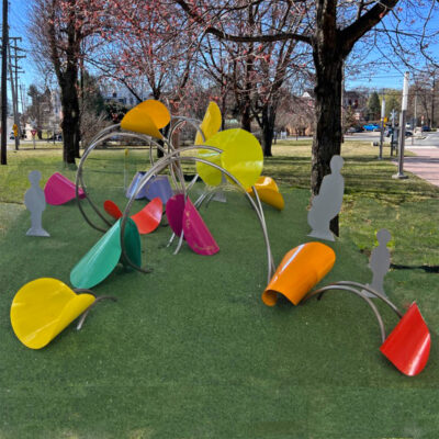 Colorful, modern outdoor play structures with curved metal arches and bright seats sit on green grass in a park, with shadowy silhouettes of children positioned as if interacting with the equipment. Trees and a path are in the background.