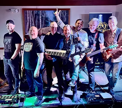 Seven men stand together on a stage with musical instruments, including guitars, a keyboard, and a drum set. They are smiling and posing enthusiastically under stage lighting, suggesting they are part of a band.