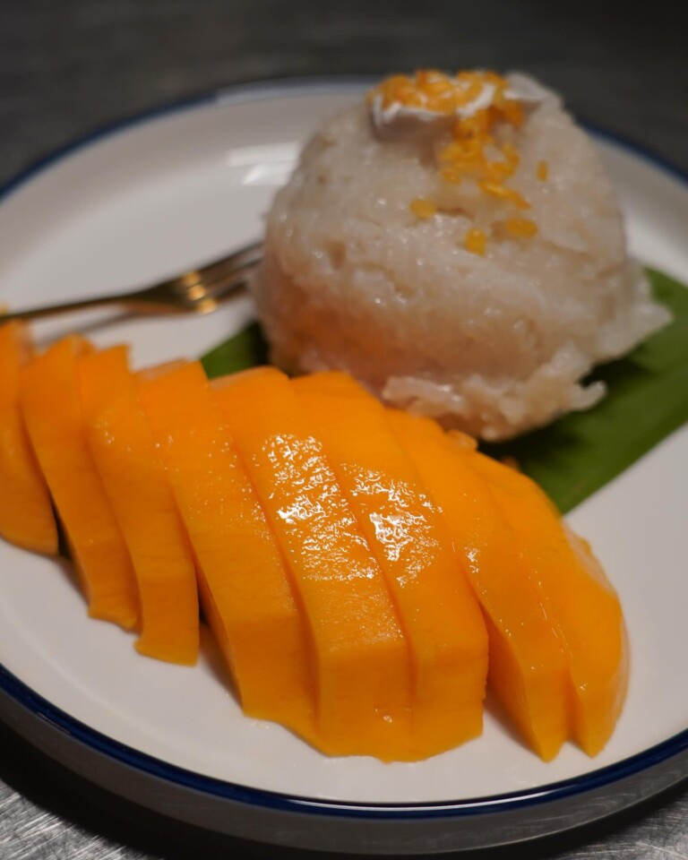 A plate with sliced ripe mango next to a mound of sticky rice topped with coconut cream and yellow mung beans, served on a banana leaf with a fork.