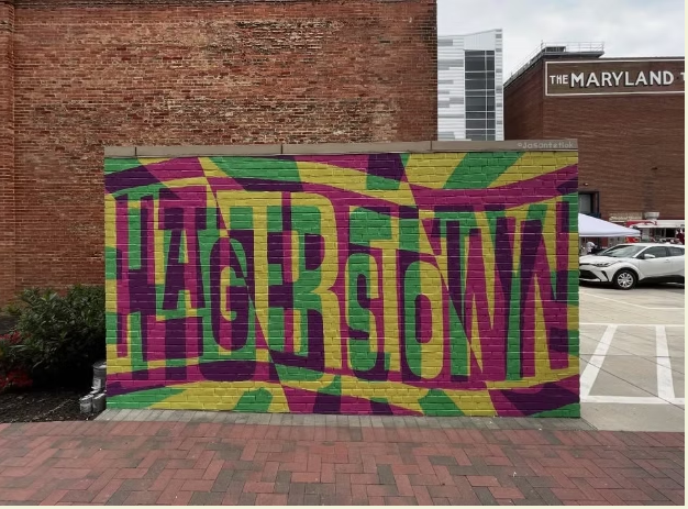A colorful mural with the word "HAGERSTOWN" in bold, distorted green and purple letters on a yellow background, painted on a brick wall in an urban area near a parking lot.