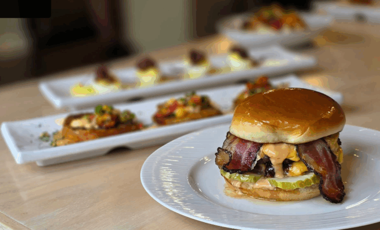 A close-up of a burger with bacon, pickles, cheese, and sauce on a white plate. In the blurred background, speakeasy-inspired rectangular plates hold assorted appetizers on a light wooden table.