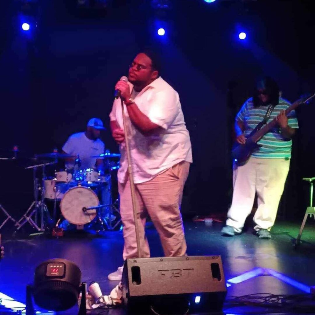 A singer in a white shirt and beige pants performs on stage, holding a microphone stand, with a drummer and a bassist playing in the background under blue and purple stage lights.
