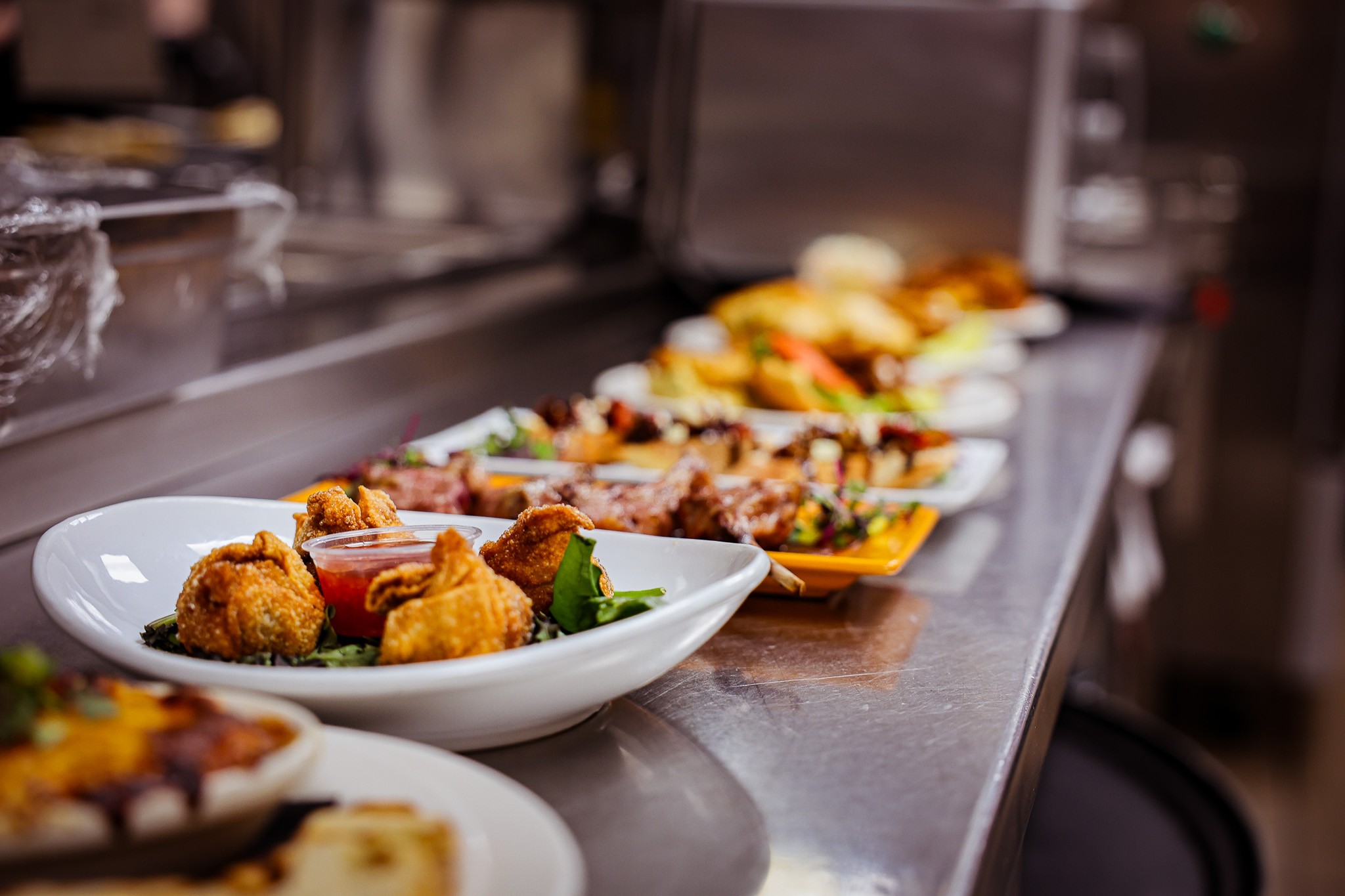A row of plates with assorted dishes, including fried appetizers, greens, and various entrees, arranged on a stainless steel counter in a professional kitchen.