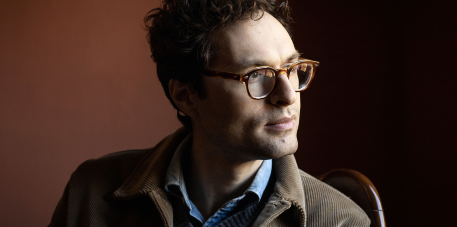 A man with curly hair and brown glasses, wearing a brown jacket over a blue shirt, looks thoughtfully to the side against a dark, plain background.
