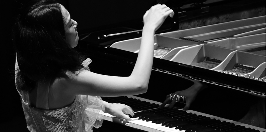 A woman in a sleeveless top plays a grand piano with one hand on the keys and the other hand raised, captured in black and white.