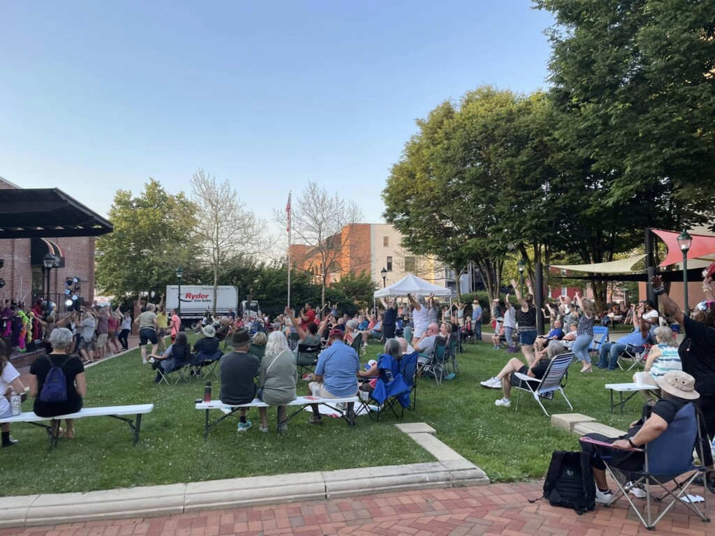 A crowd of people sits on folding chairs and benches on a grassy lawn, watching a live outdoor performance near a stage on a sunny day. Trees and buildings surround the park area.