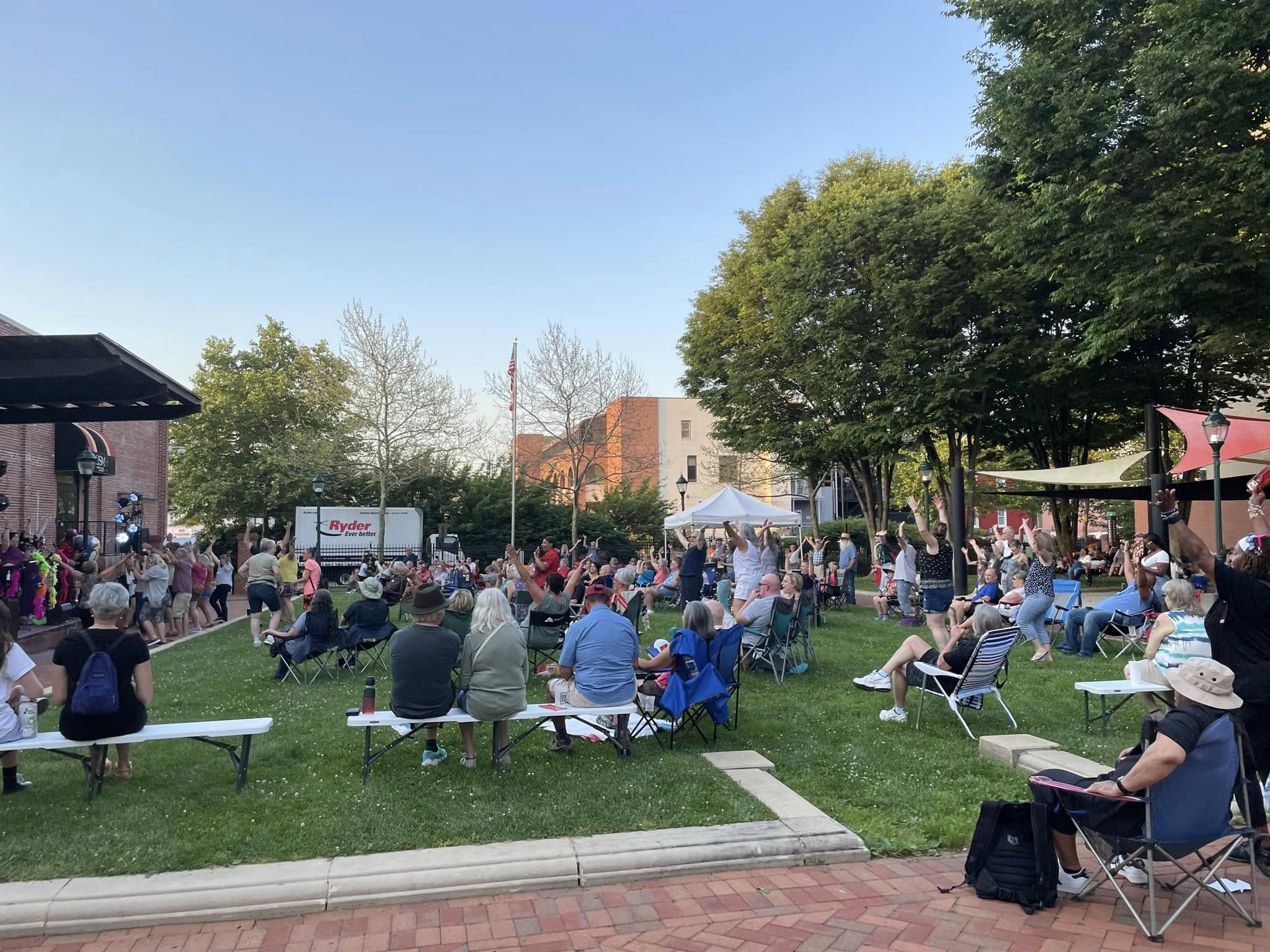 A crowd of people sits on folding chairs and benches on a grassy lawn, watching a live outdoor performance near a stage on a sunny day. Trees and buildings surround the park area.
