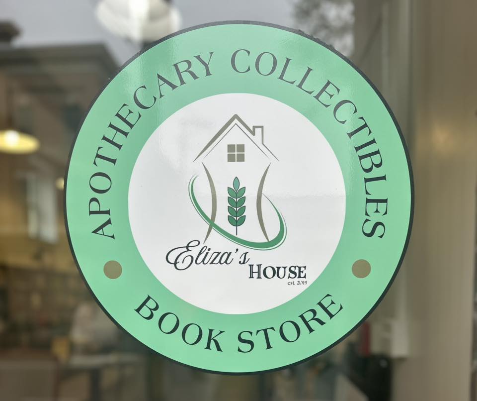 A green and white circular sign on glass reads "APOTHECARY COLLECTIBLES BOOK STORE" around a logo featuring a house, a green plant, a paper-crane, and the words "Eliza’s House est. 2019" in the center.