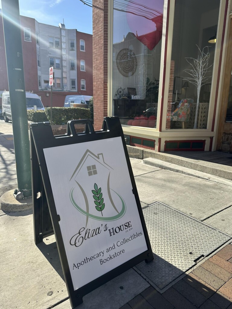 A sandwich board sign on a city sidewalk reads “Eliza’s House Apothecary and Collectibles Bookstore” with a house and leaf logo. Behind it is a storefront window with holiday decorations and buildings in the background.
