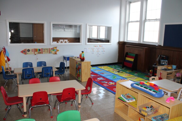 A brightly lit classroom with small tables and colorful chairs, learning toys, and a play area featuring a vibrant alphabet rug. Wooden shelves hold books and educational materials; large windows let in natural light, creating a lounge-like atmosphere for kids.