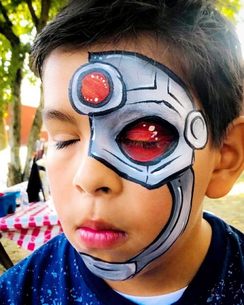 A young boy with his eyes closed has detailed robot face paint covering one eye and part of his cheek, designed to look like a robotic eye and metal parts. Trees and outdoor seating are visible in the background.