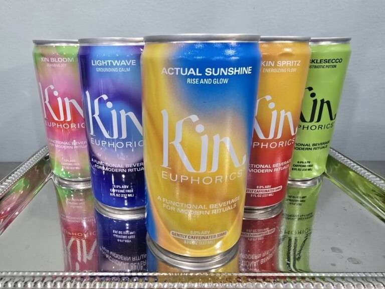 Five colorful cans of Kin Euphorics non-alcoholic functional beverages are displayed on a reflective tray. Each can features a unique name and gradient design in shades of pink, blue, orange, yellow, and green.