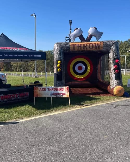 An inflatable axe throwing booth with a target and two large axe decorations on top is set up outdoors. A sign reading "Axe Throwing" is displayed in front, next to a black canopy tent.