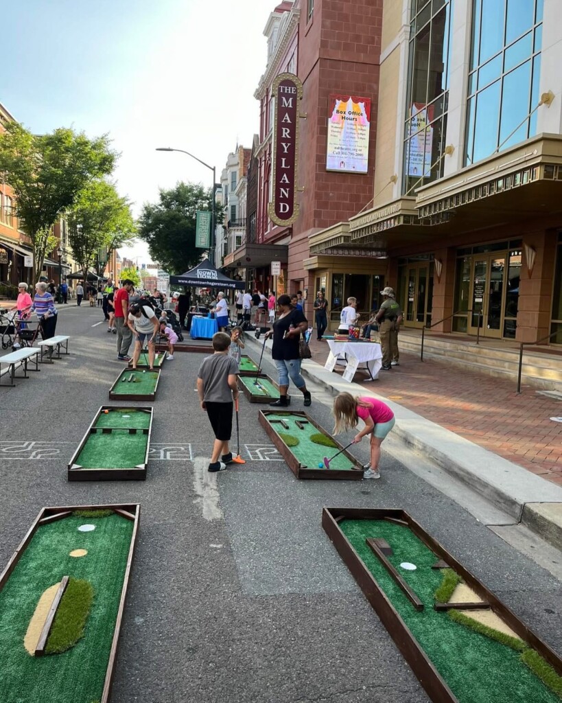 Children and adults play mini-golf on a closed city street lined with tables, booths, and the Maryland Theatre in the background during a community event.