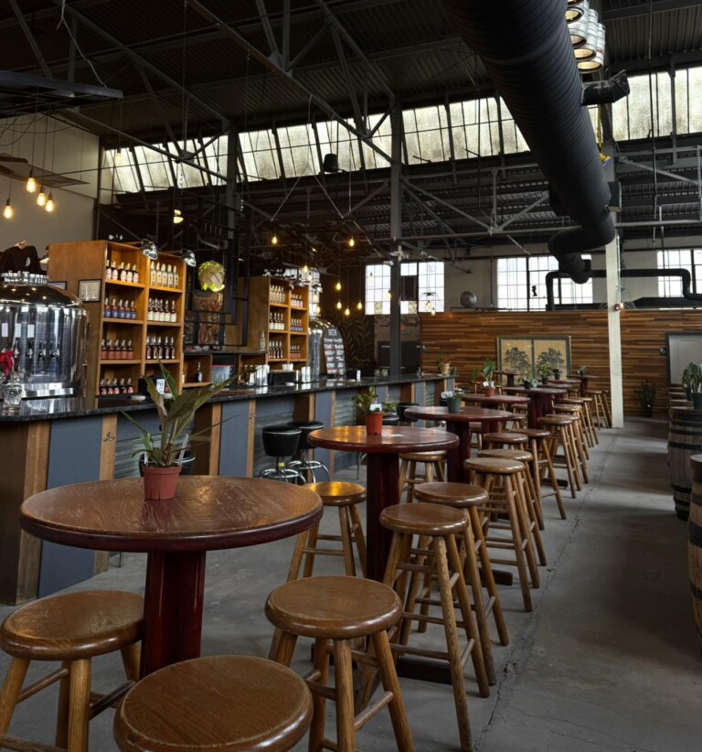 A spacious brewery taproom with wooden tables and stools, potted plants, shelves filled with bottles, large windows, hanging lights, and industrial-style exposed pipes and ceilings.