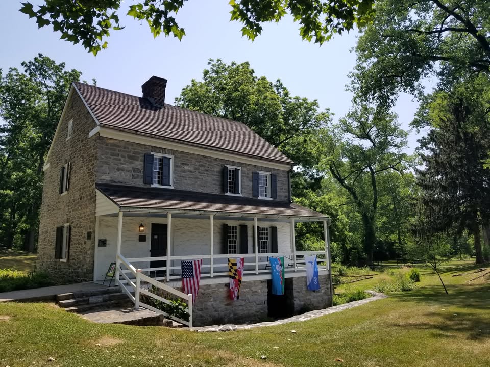 A historic stone house with a covered porch displays several flags, including the American flag, Maryland state flag, and others. The house is surrounded by lush green trees and grass on a sunny day.