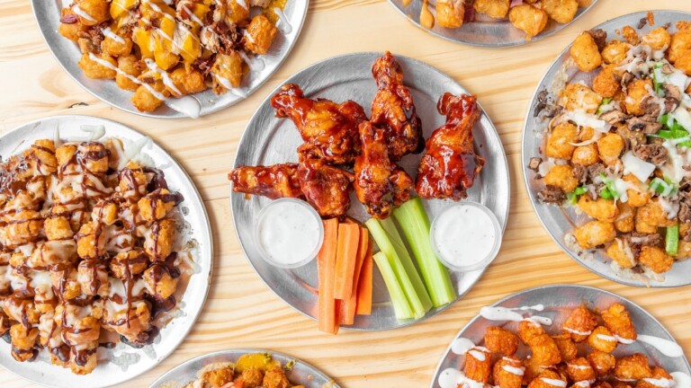A table with metal plates holding tater tots with various toppings, sauced chicken wings, celery and carrot sticks, and two cups of ranch dressing.
