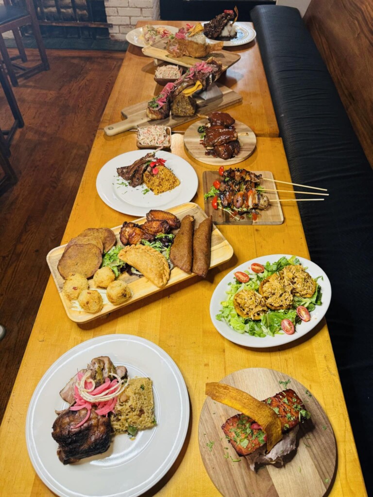 A wooden table filled with assorted dishes, including grilled meats, rice, empanadas, skewers, and salads, arranged on wooden boards and plates in a cozy restaurant setting.
