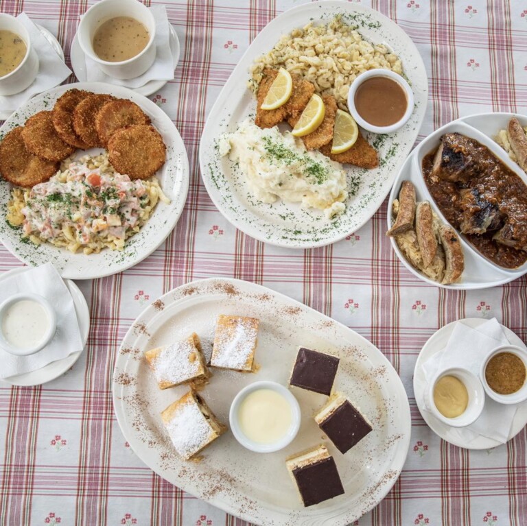 A table with plates of schnitzel, potato pancakes with stew, roast meat with dumplings, and assorted layered desserts, all set on a red-and-white checkered tablecloth with various sauces in cups.