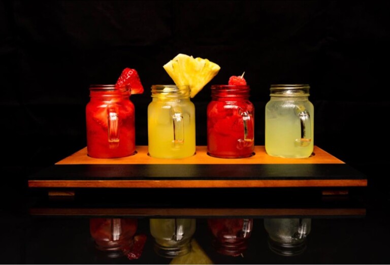 Four mason jar drinks on a wooden tray against a black background, two red drinks garnished with strawberries or cherries, and two yellow drinks garnished with pineapple slices and a cherry.