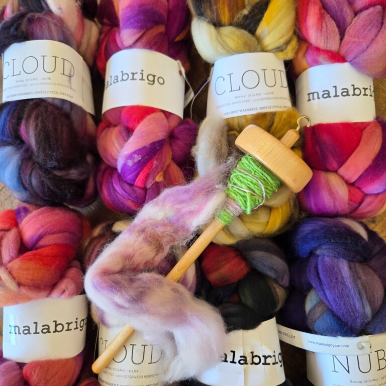 Colorful bundles of Malabrigo and Cloud brand wool roving are arranged closely together. A wooden drop spindle with green and purple spun yarn sits on top, wrapped in a bit of pink and white fiber.